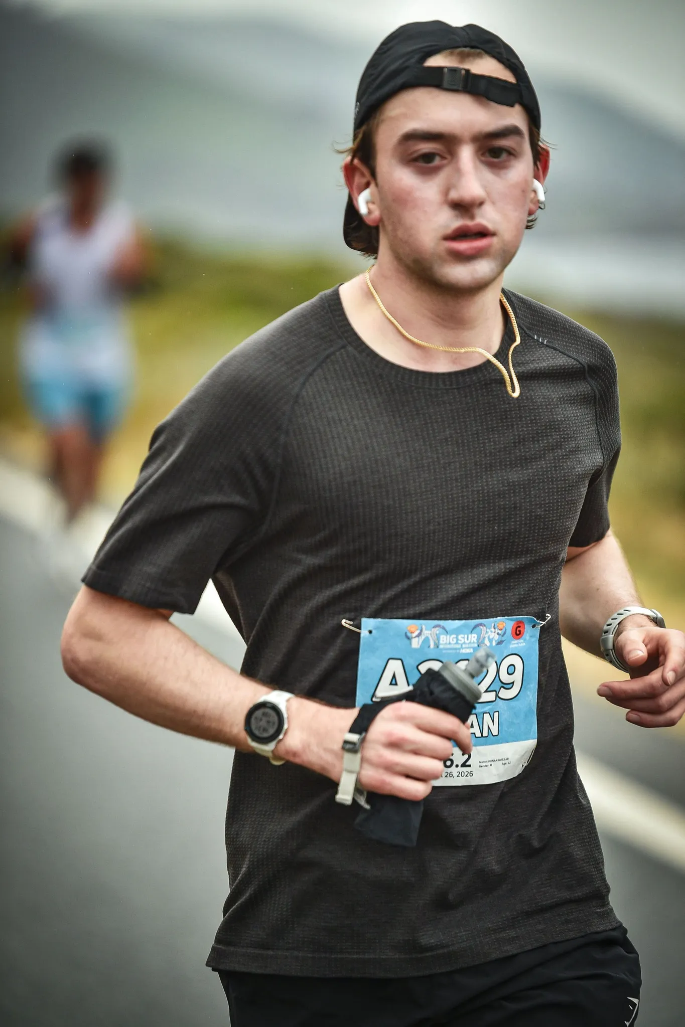 Close-up of Ronan mid-stride in a black T-shirt and backwards cap, race bib A2129 pinned on, a blurred runner behind.