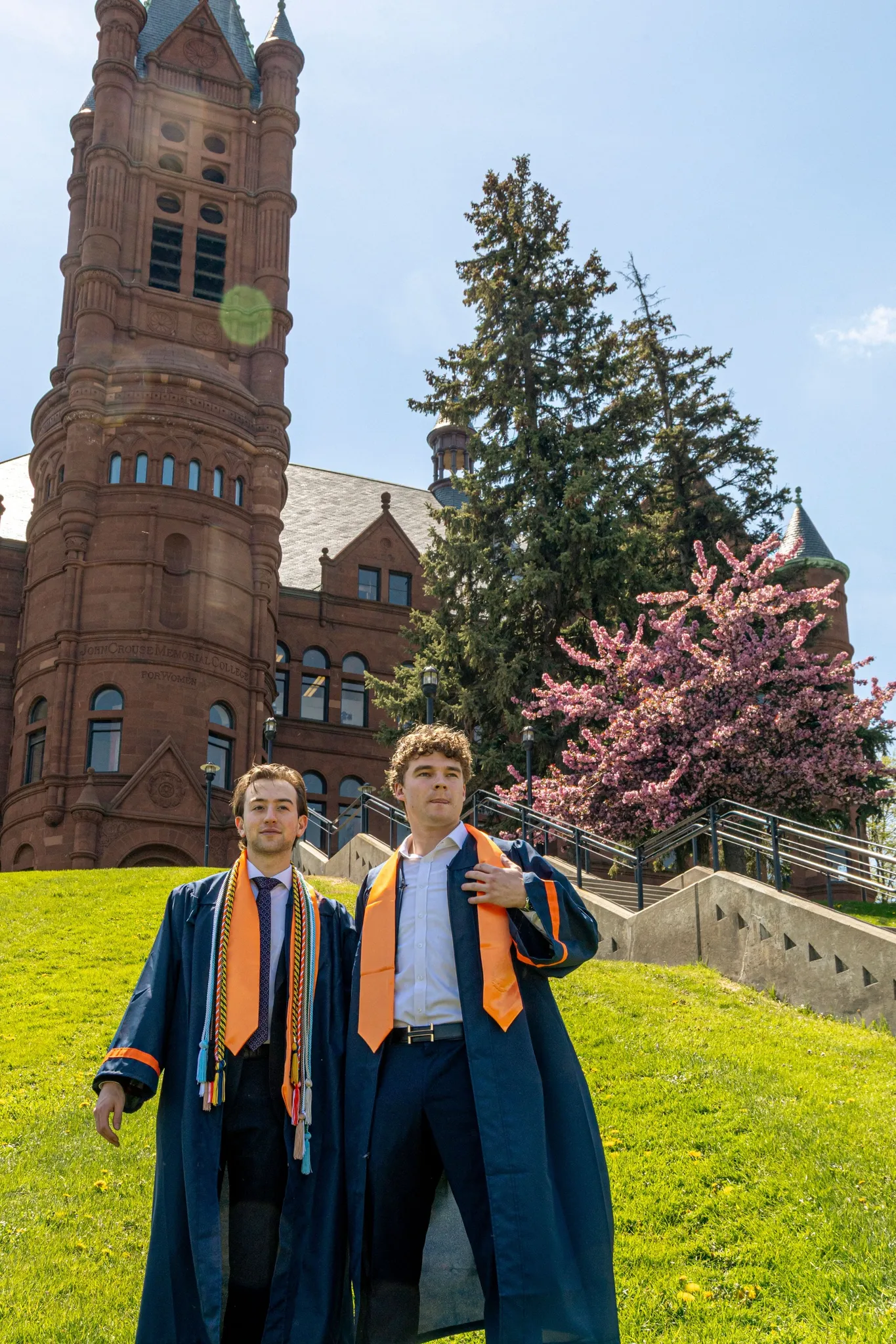 Ronan and a friend in navy gowns with orange stoles on a steep green lawn below a red sandstone clock tower and a pink blossoming tree.