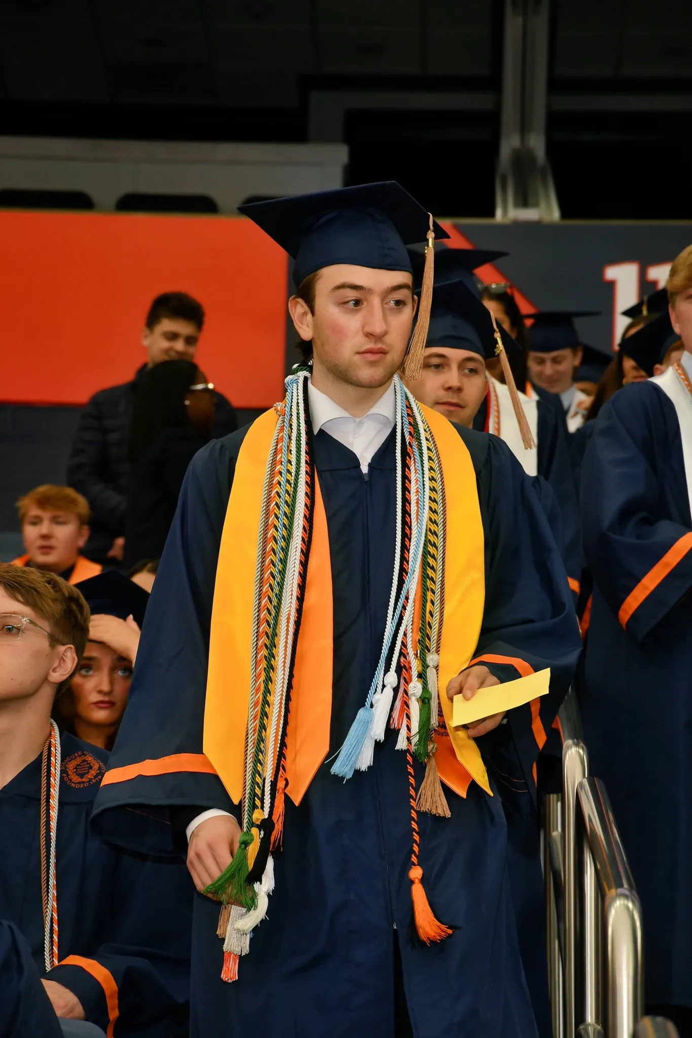 Ronan in a navy cap and gown walking toward the camera holding a diploma cover, graduates and the stadium behind him.