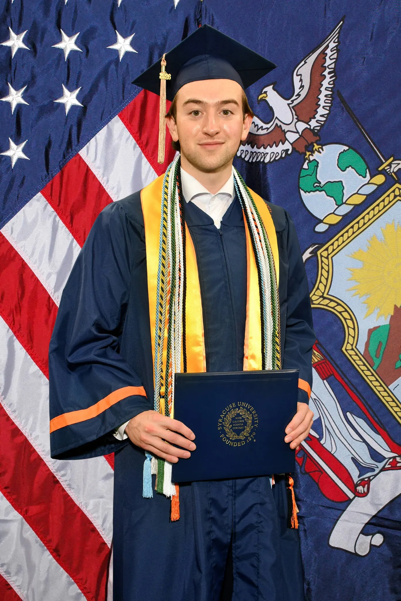 Ronan in a navy cap and gown with stoles, holding a diploma cover in front of the American and New York State flags.