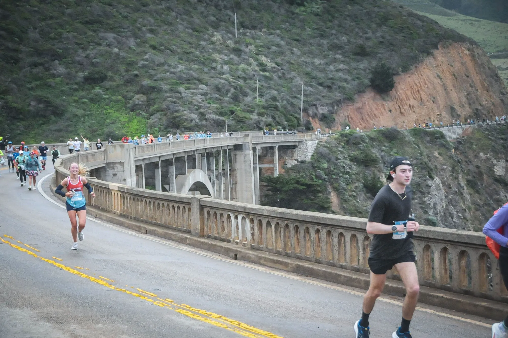 Ronan running across a concrete arch bridge on a coastal highway, cliffs and scrub behind him and other runners strung out along the road.