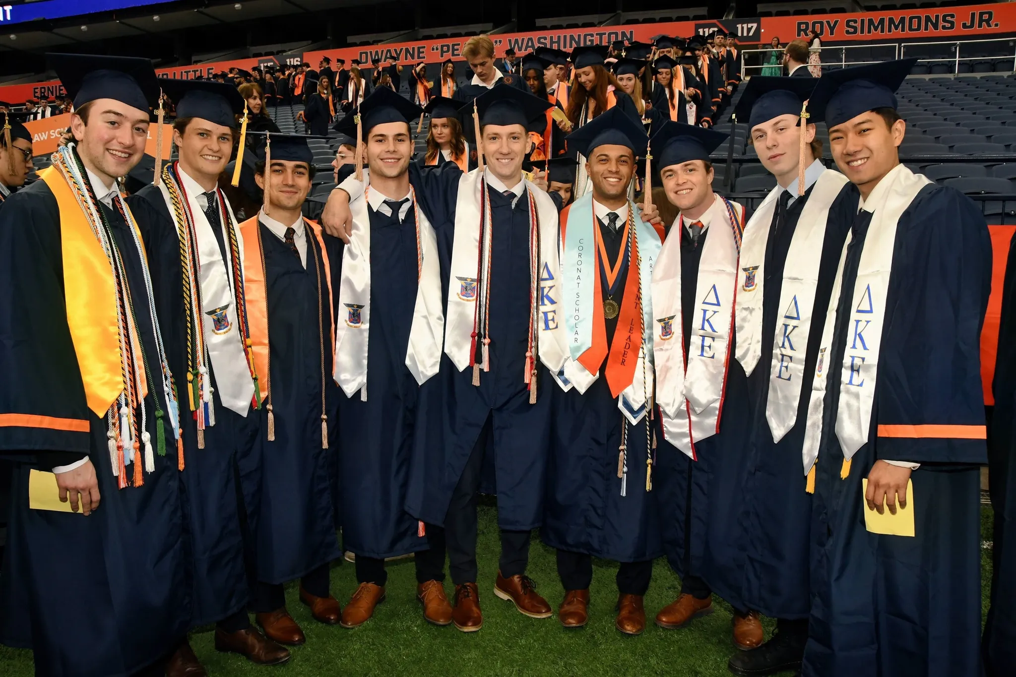 A group of about a dozen graduates in navy gowns and stoles standing together on the stadium floor.