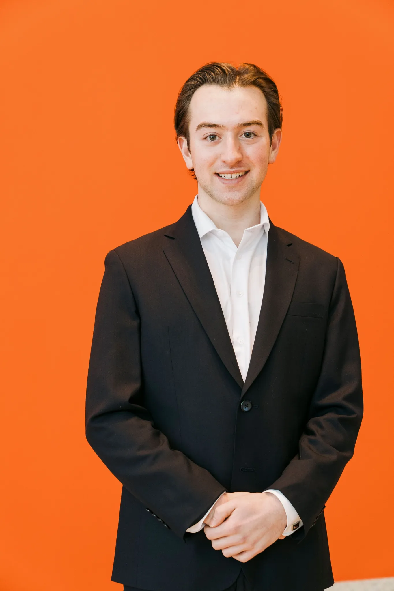 Ronan in a dark suit and white shirt, hands clasped, standing in front of a bright orange wall.