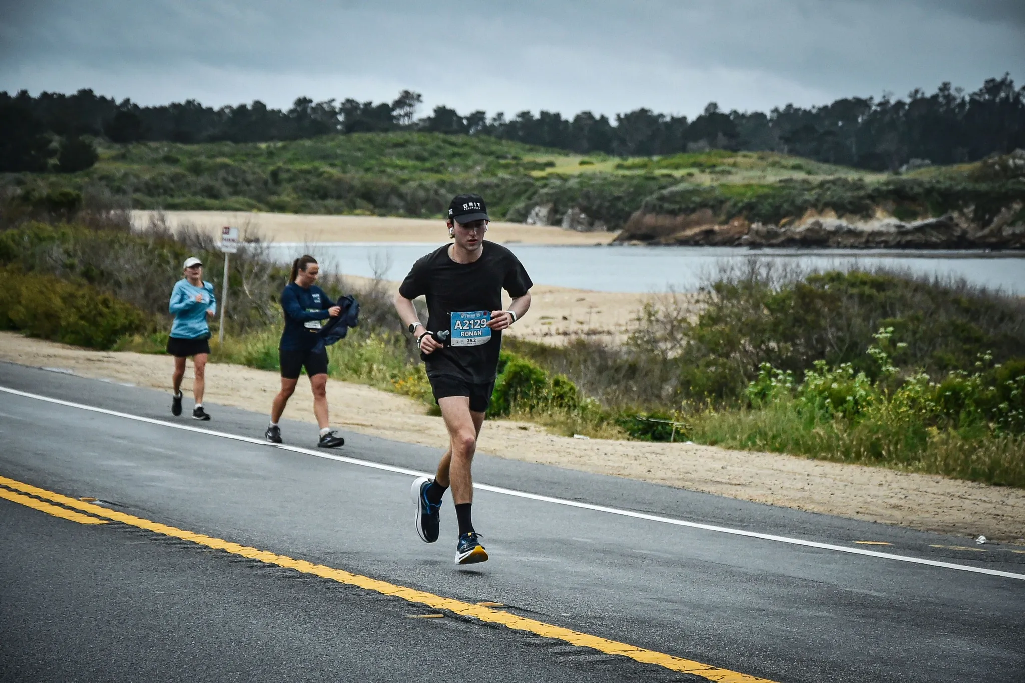 Ronan running along a two-lane coastal road beside a lagoon and beach, two other runners behind him.