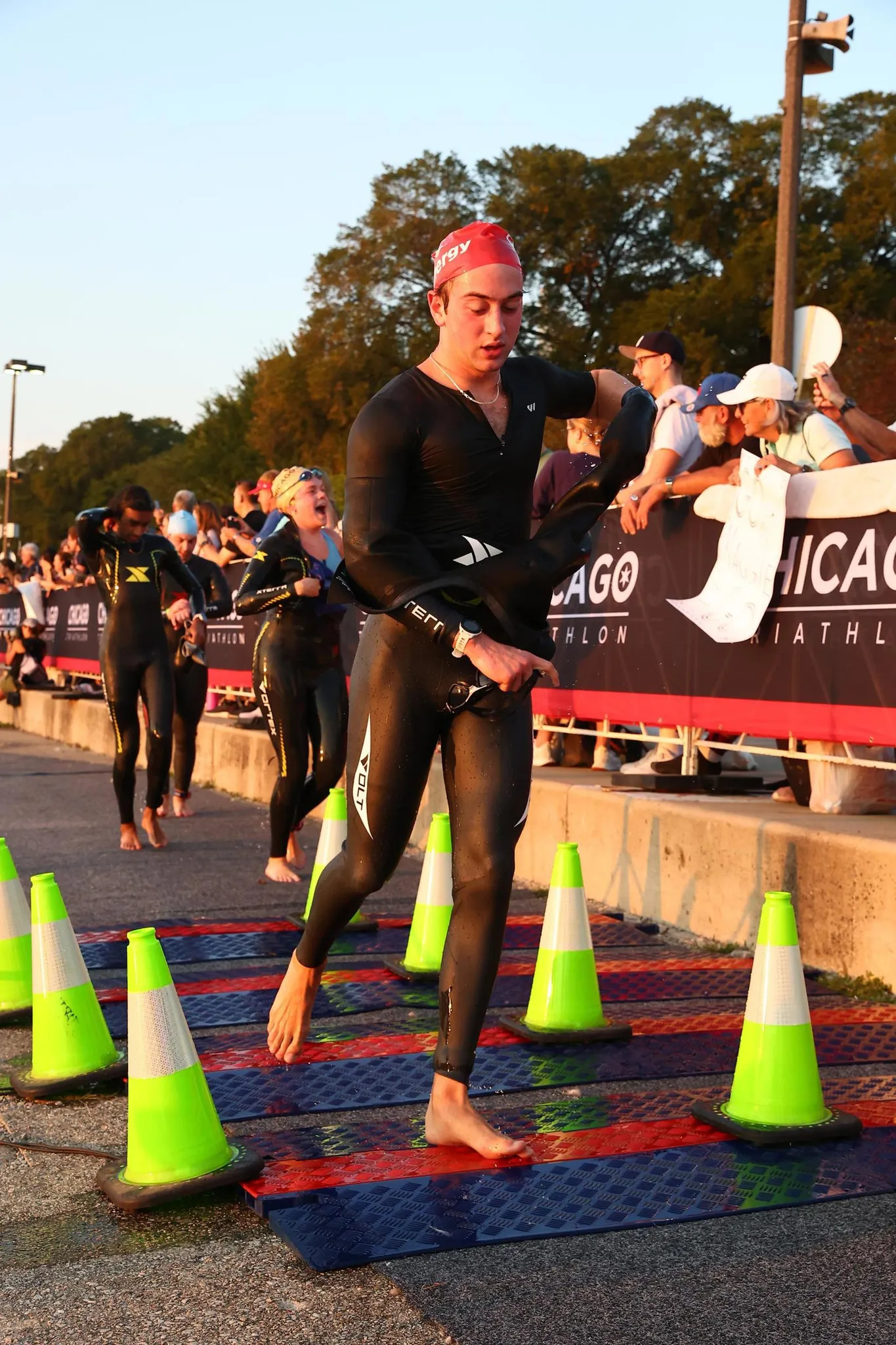 Ronan in a black wetsuit and red swim cap running barefoot out of the swim exit between green cones, spectators along the barrier.