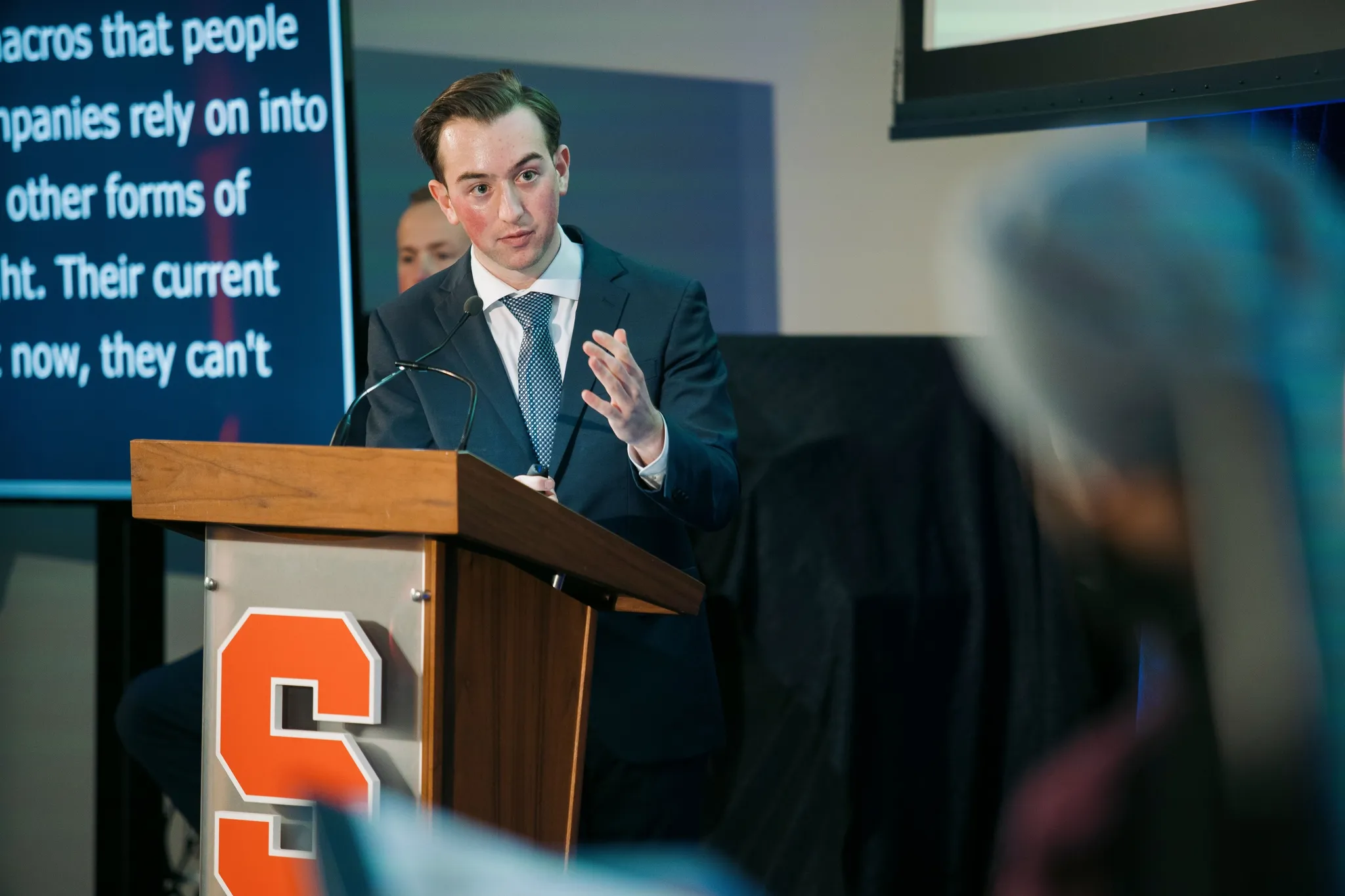 Ronan in a suit gesturing as he speaks at a Syracuse podium, a slide of text on the screen behind him and audience members in the foreground.