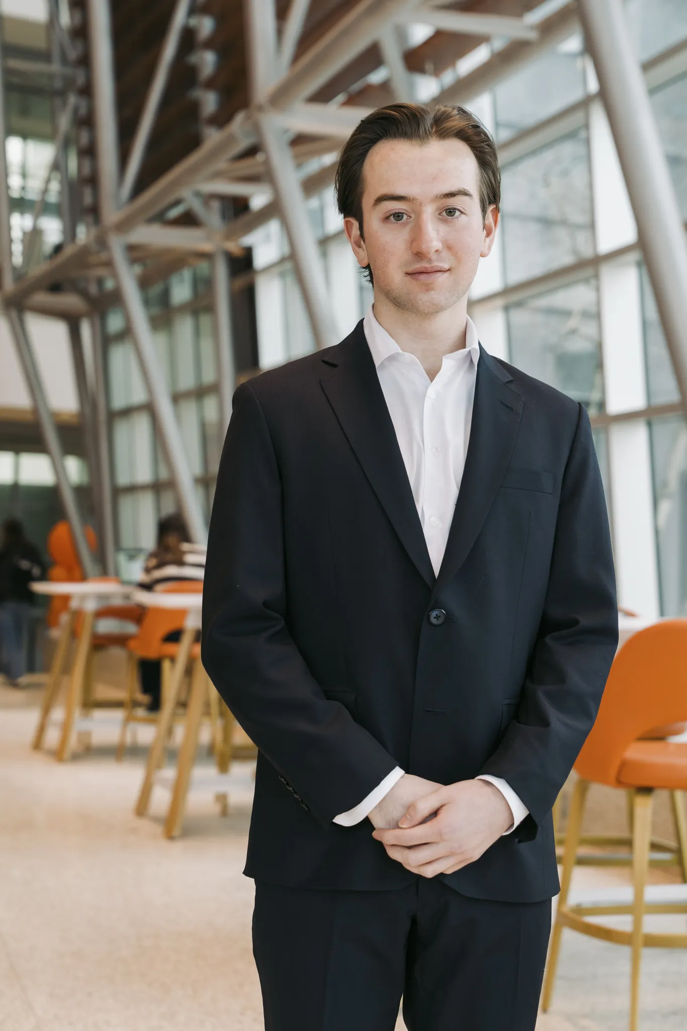 Full-length portrait of Ronan in a dark suit and white shirt, hands clasped, in a bright atrium with orange chairs behind.