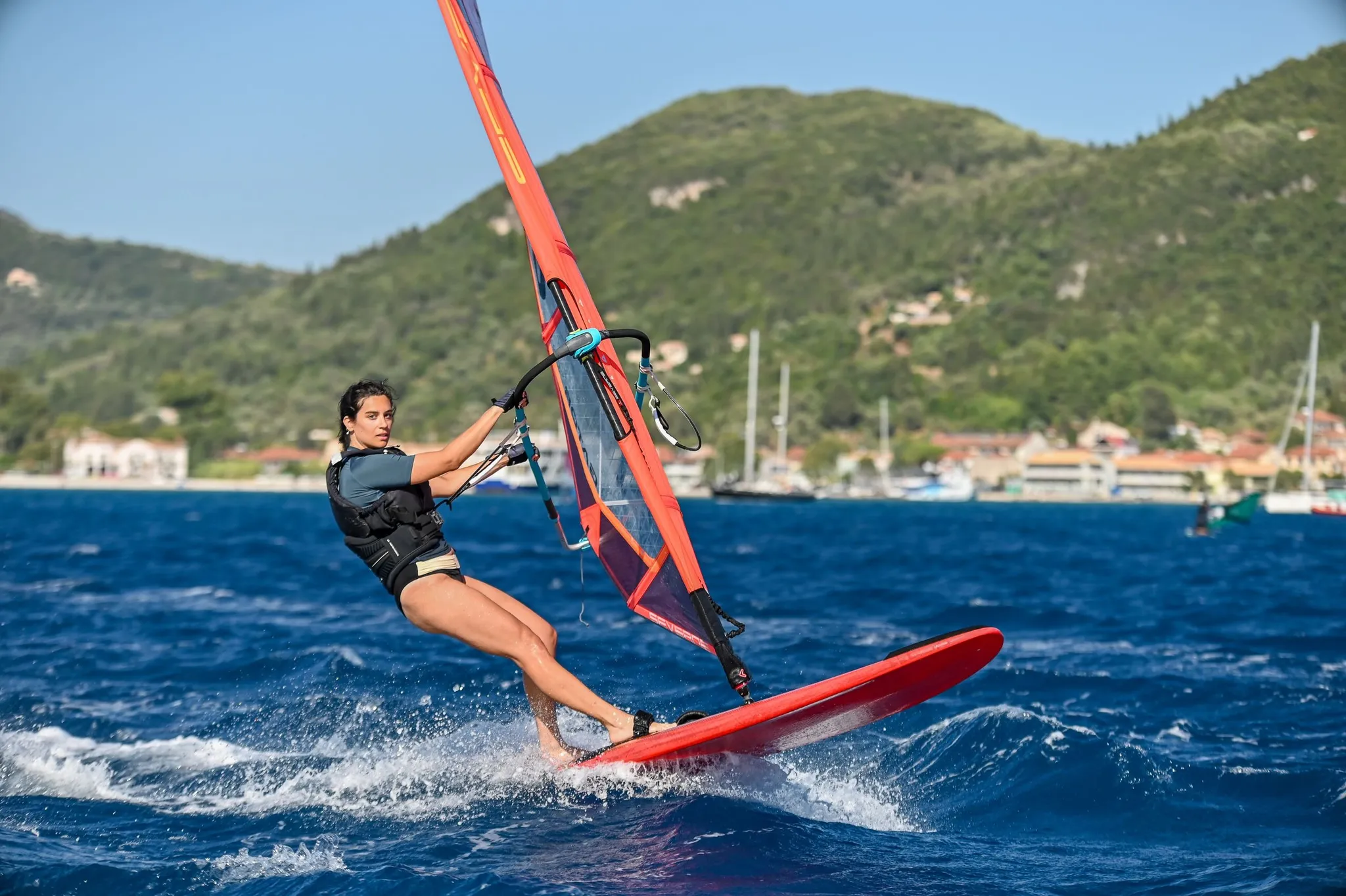 Ronan leaning far back off a red windsurfer, spray behind the board, green hills and moored boats in the background.