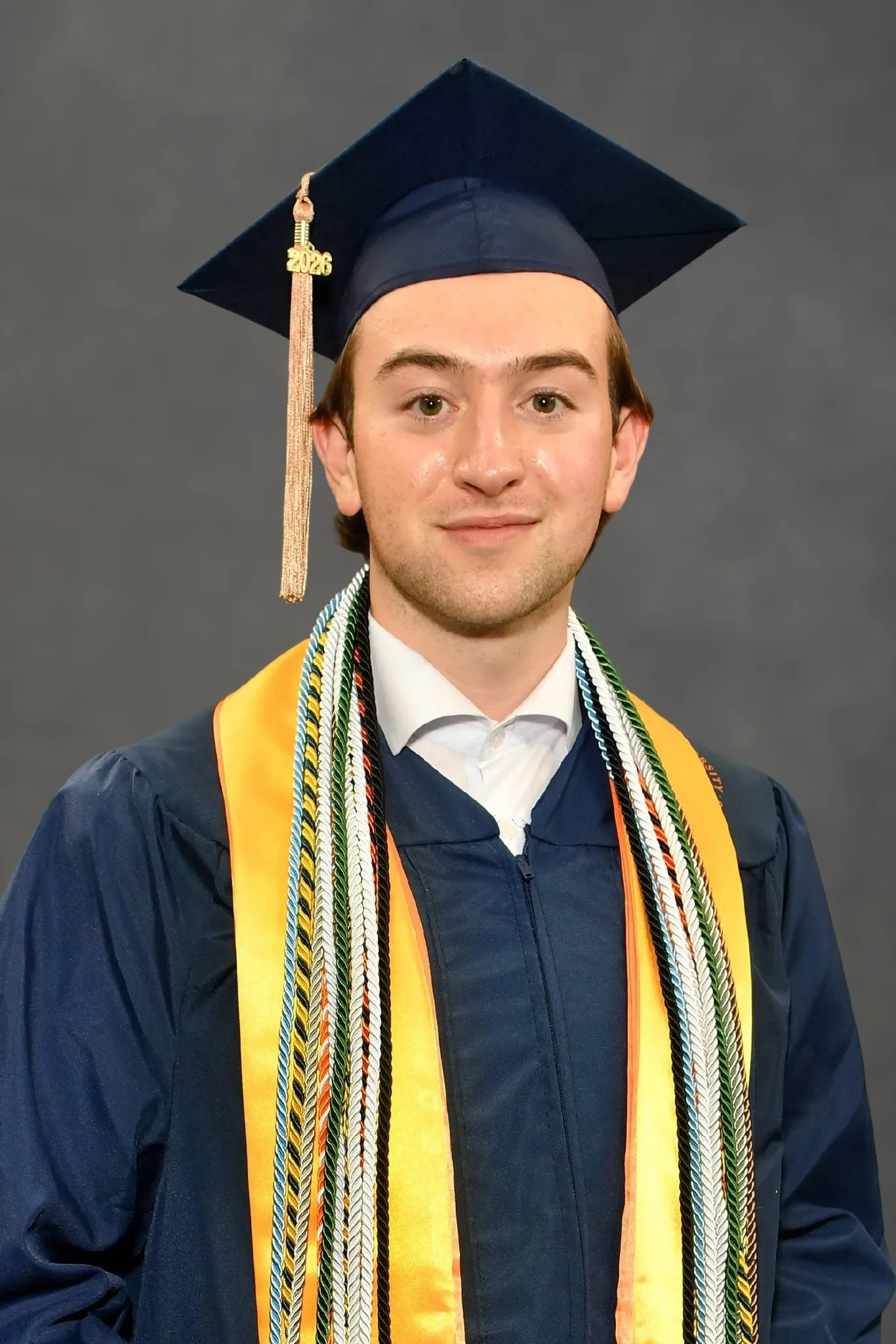 Studio portrait of Ronan smiling in a navy cap and gown with gold and green stoles, against a grey backdrop.