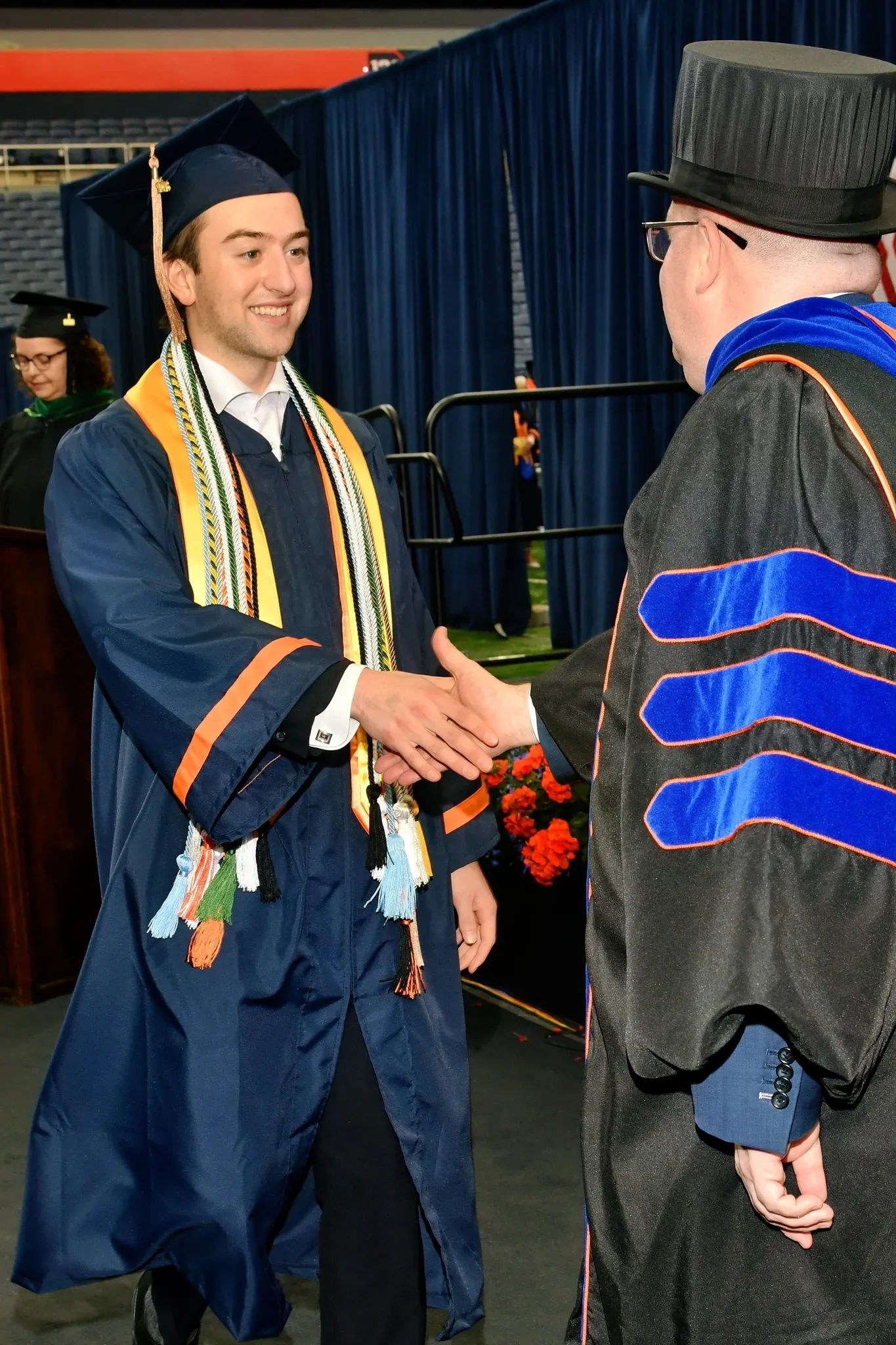 Ronan in a navy gown and mortarboard with gold stoles, smiling and shaking hands on stage with a faculty member in a top hat and doctoral robes.