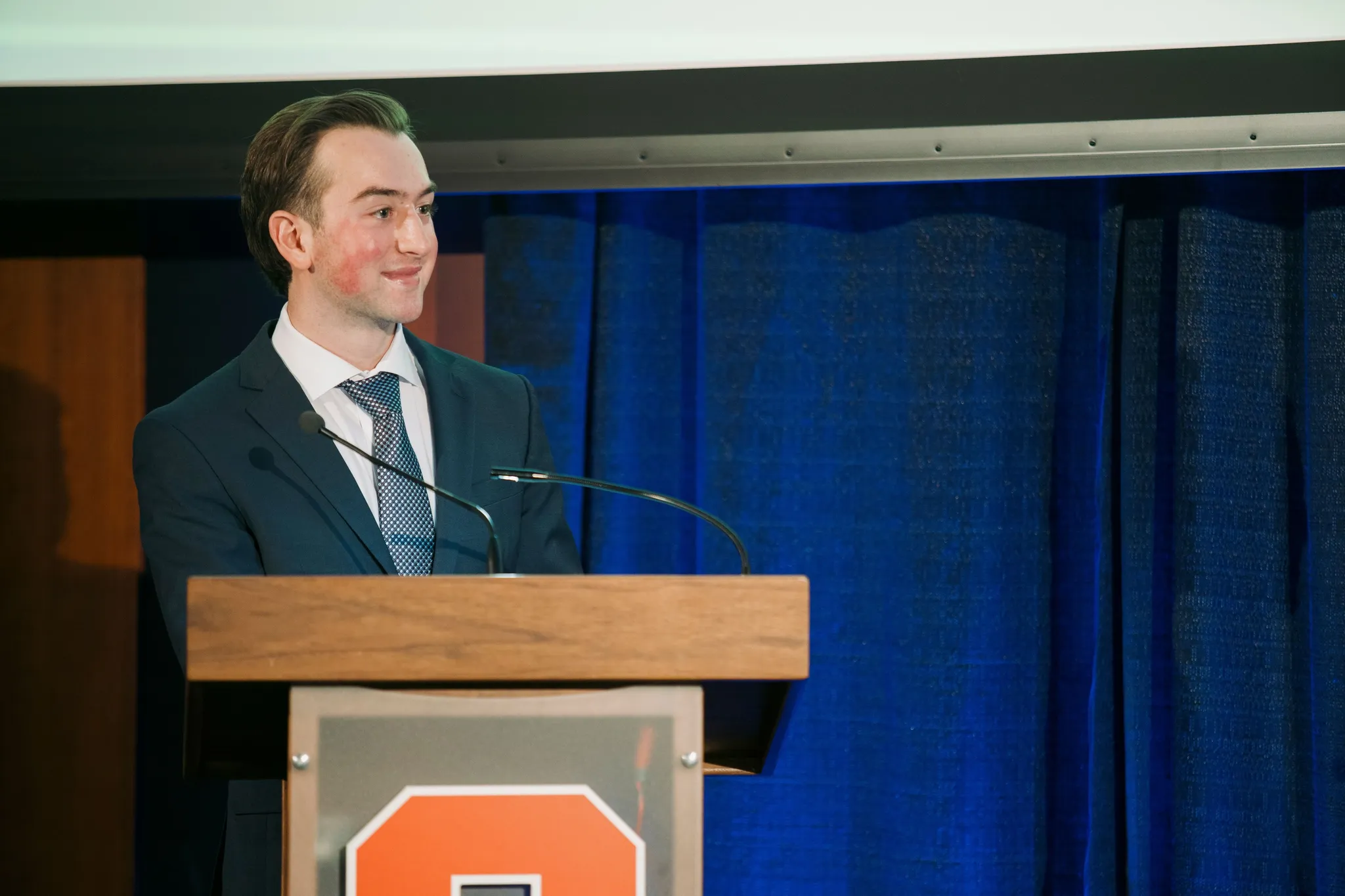 Ronan smiling at a wooden podium with the Syracuse S on its front, a blue curtain behind.