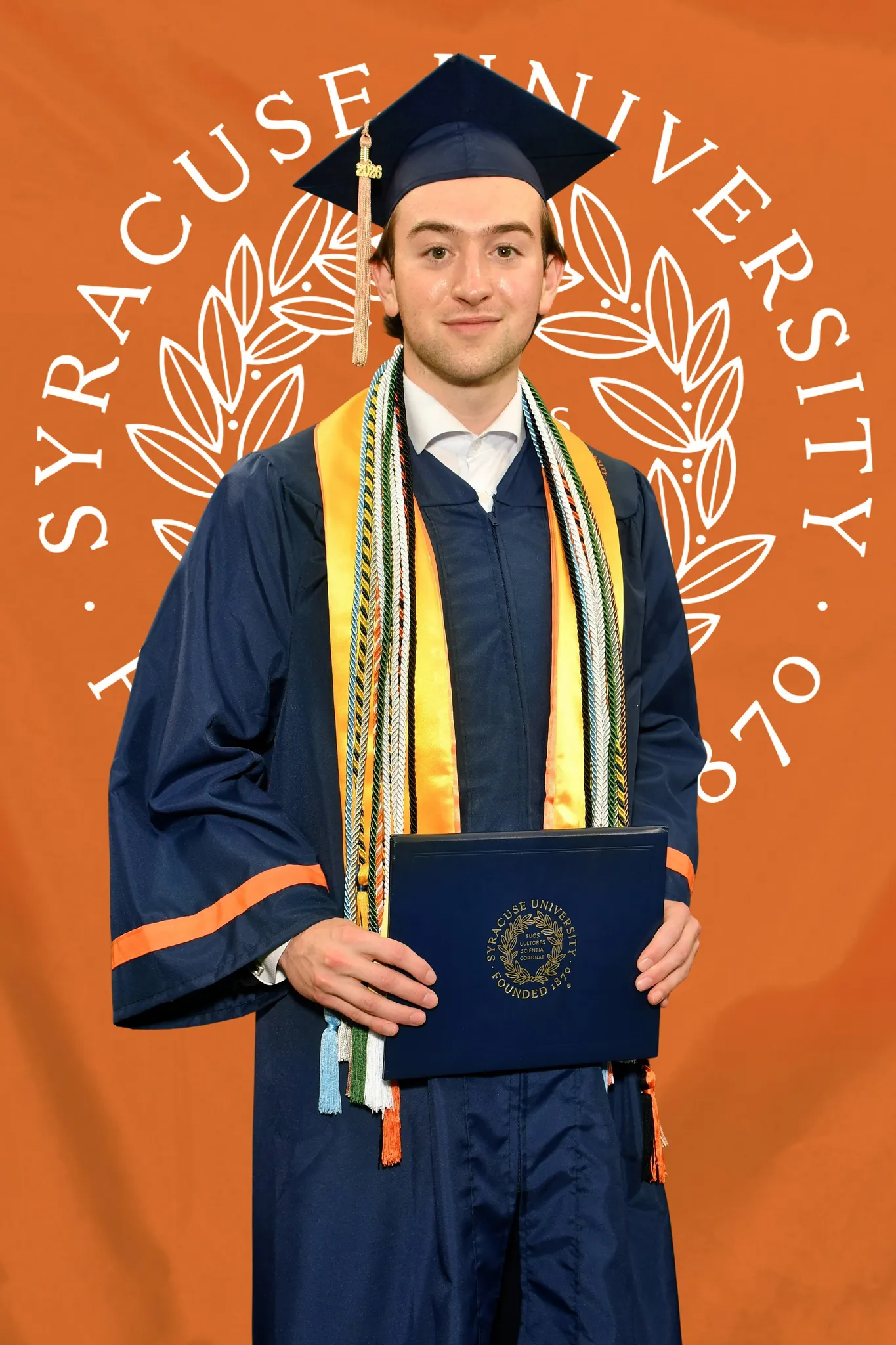 Ronan in a navy cap and gown holding a diploma cover in front of a bright orange backdrop printed with the Syracuse University seal.
